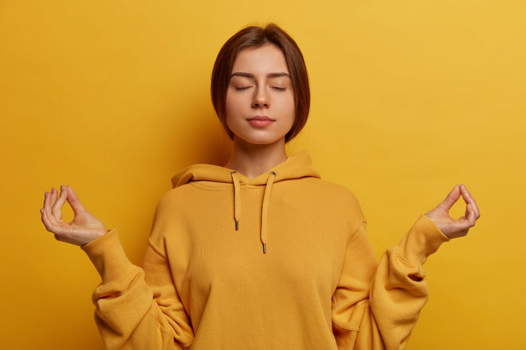 Prise de vue horizontale d’une femme patiente qui est sur l’auto-isolement à la maison, se détend avec le yoga et médite à l’intérieur, garde les mains dans le geste zen, atteint nirvana, respire profondément, porte un sweat-shirt jaune