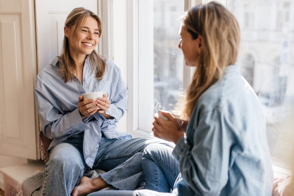 two-beautiful-caucasian-women-mother-daughter-are-sitting-windowsill-drinking-tea-talking-each-other-relationships-joint-time-concept